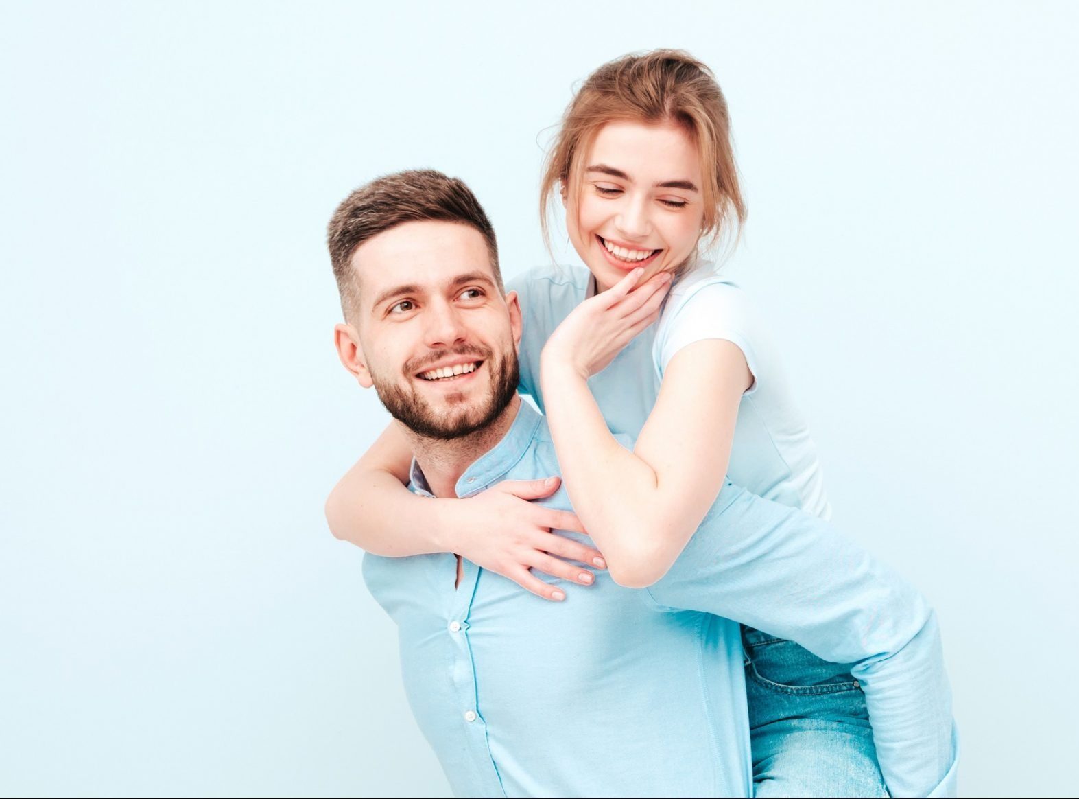 Smiling beautiful woman and her handsome boyfriend.Happy cheerful family having tender moments near light blue wall in studio.Pure models hugging.Embracing each other.Male gives piggyback riding